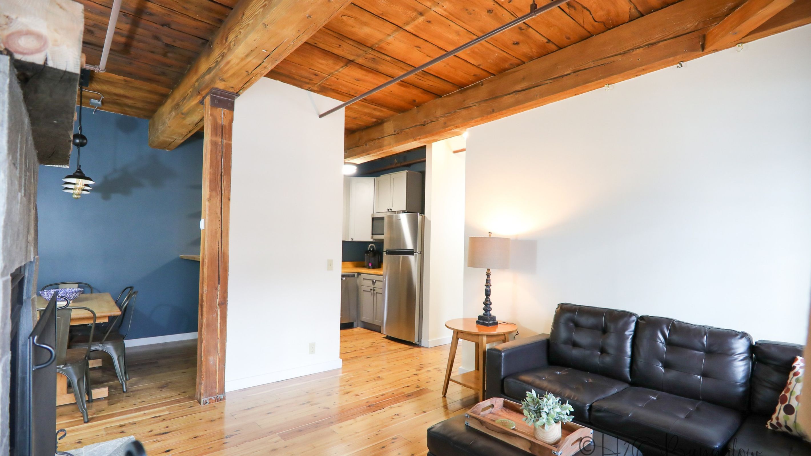 A living room with a black leather sofa, wooden flooring, a dining area with a table and chairs, and a kitchen with a refrigerator and microwave.