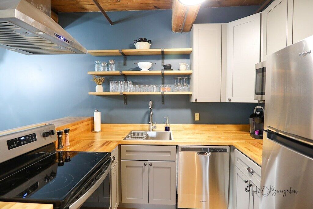 A compact kitchen with a stainless steel refrigerator, a dishwasher, a stove, and wooden shelves above the sink.