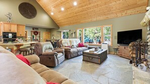 A spacious living room with a high ceiling, a fireplace, a television, and a kitchen area with wooden cabinets and a clock.