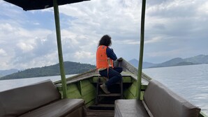 A person sitting on a bench on a boat with a view of a lake and hills in the background. - Virunga House (Muhoza)