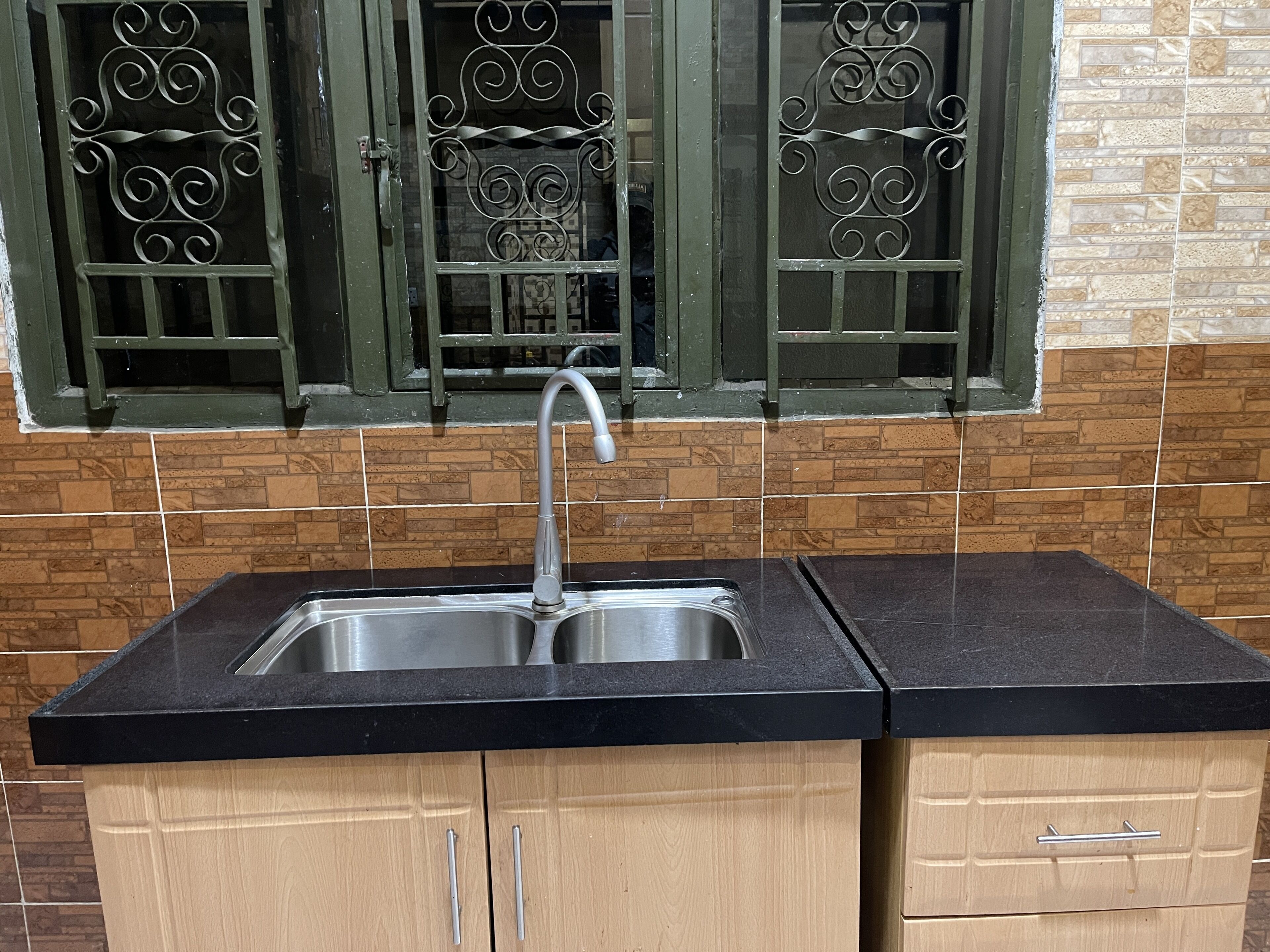 A kitchen with a black countertop, stainless steel sink, wooden cabinets, and decorative window grills.