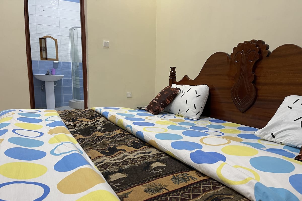 A bedroom with two beds, a wooden headboard, a patterned bedspread, a rug, and a bathroom visible in the background.