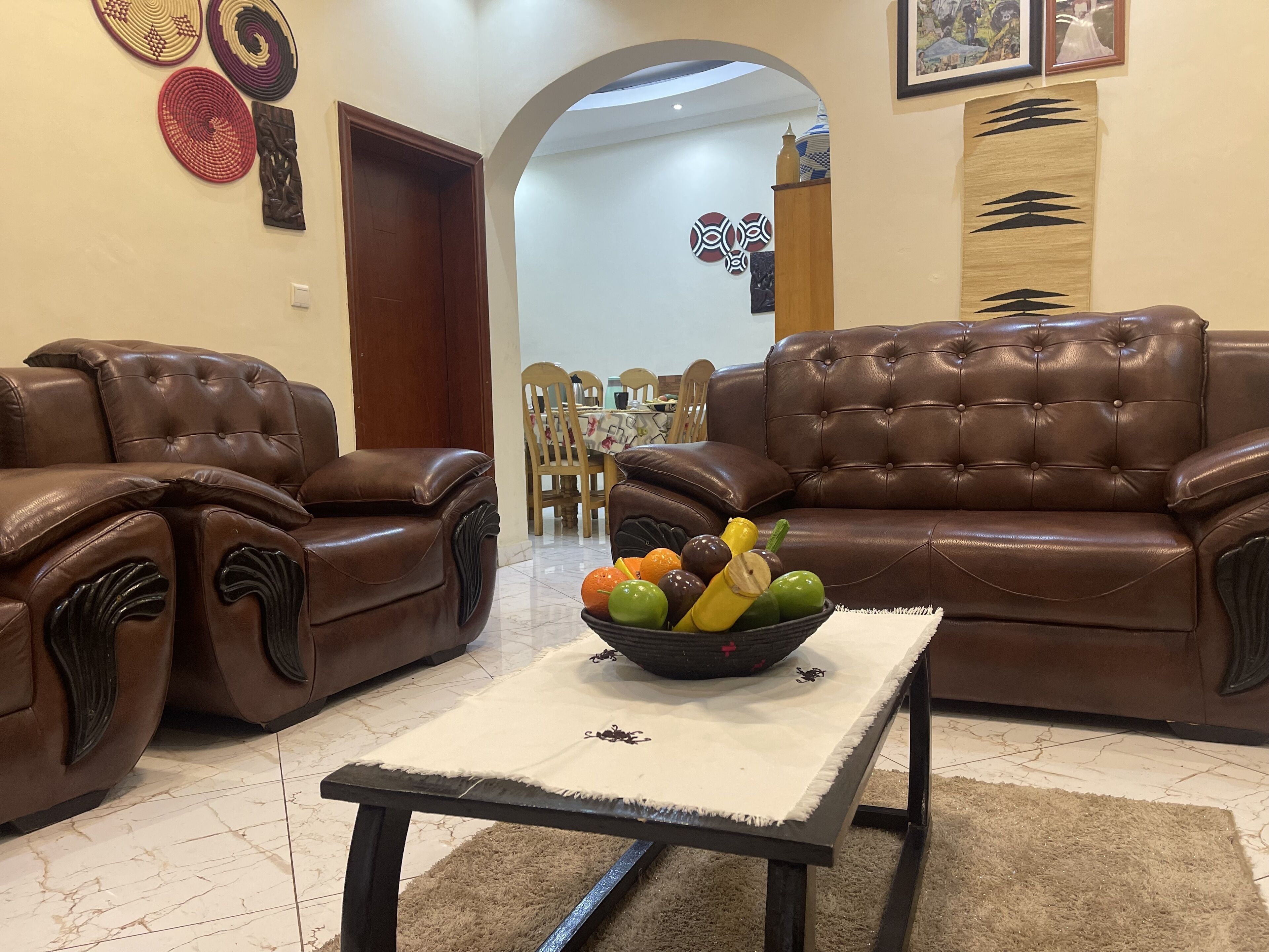 A living room with two brown leather sofas, a coffee table with a fruit bowl, and a dining area in the background.