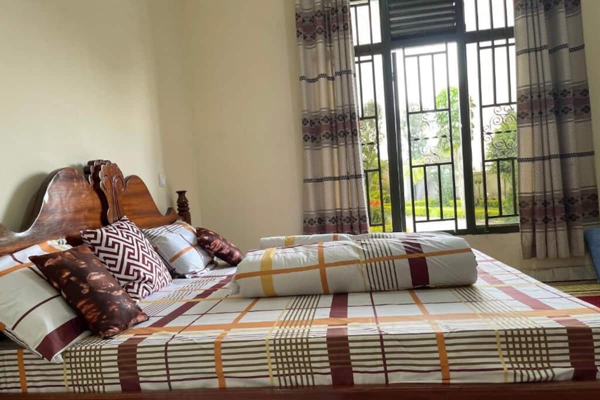 A bed with a patterned bedspread and pillows, a wooden headboard, and a view of greenery through a window with metal bars.