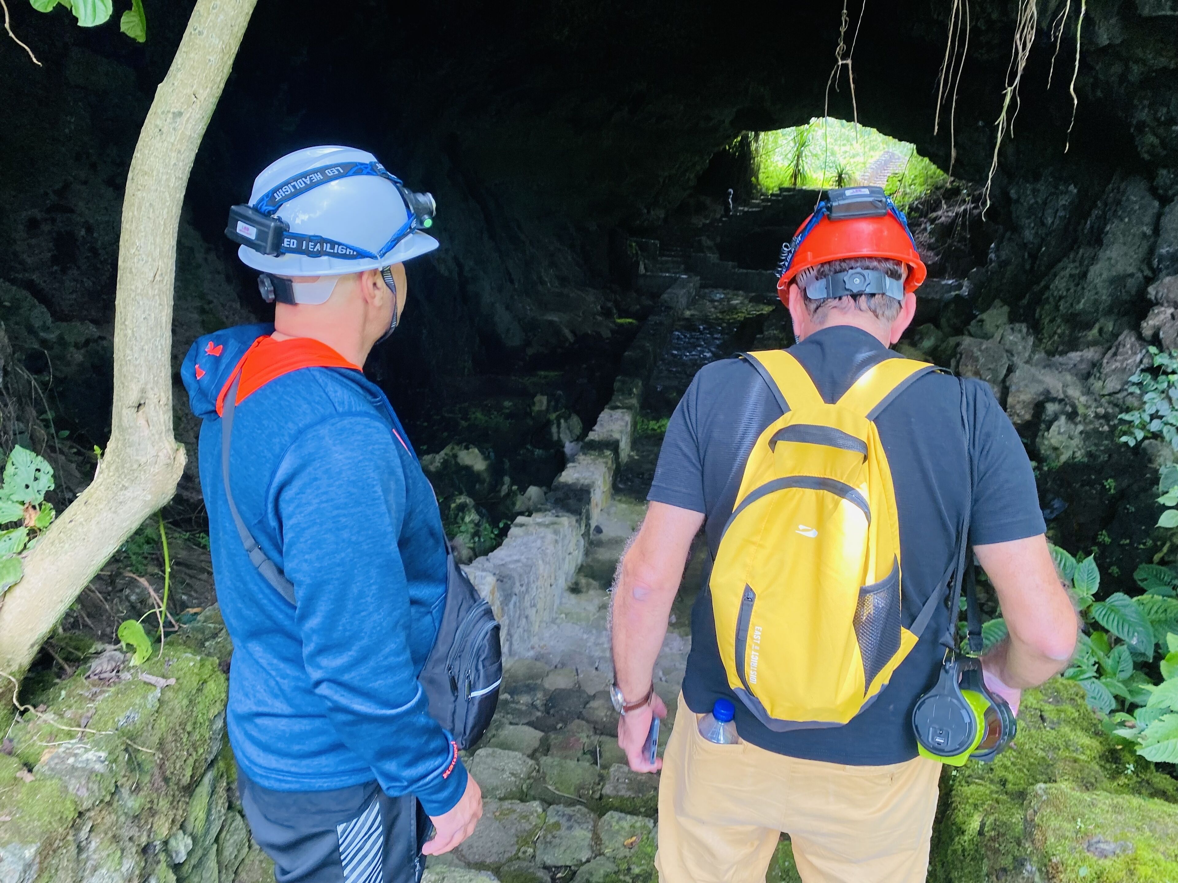 Dos personas con cascos y mochilas, de pie en la entrada de una cueva.