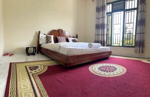 A bedroom with a wooden bed, patterned pillows, a red carpet with a circular design, and a window with metal bars.