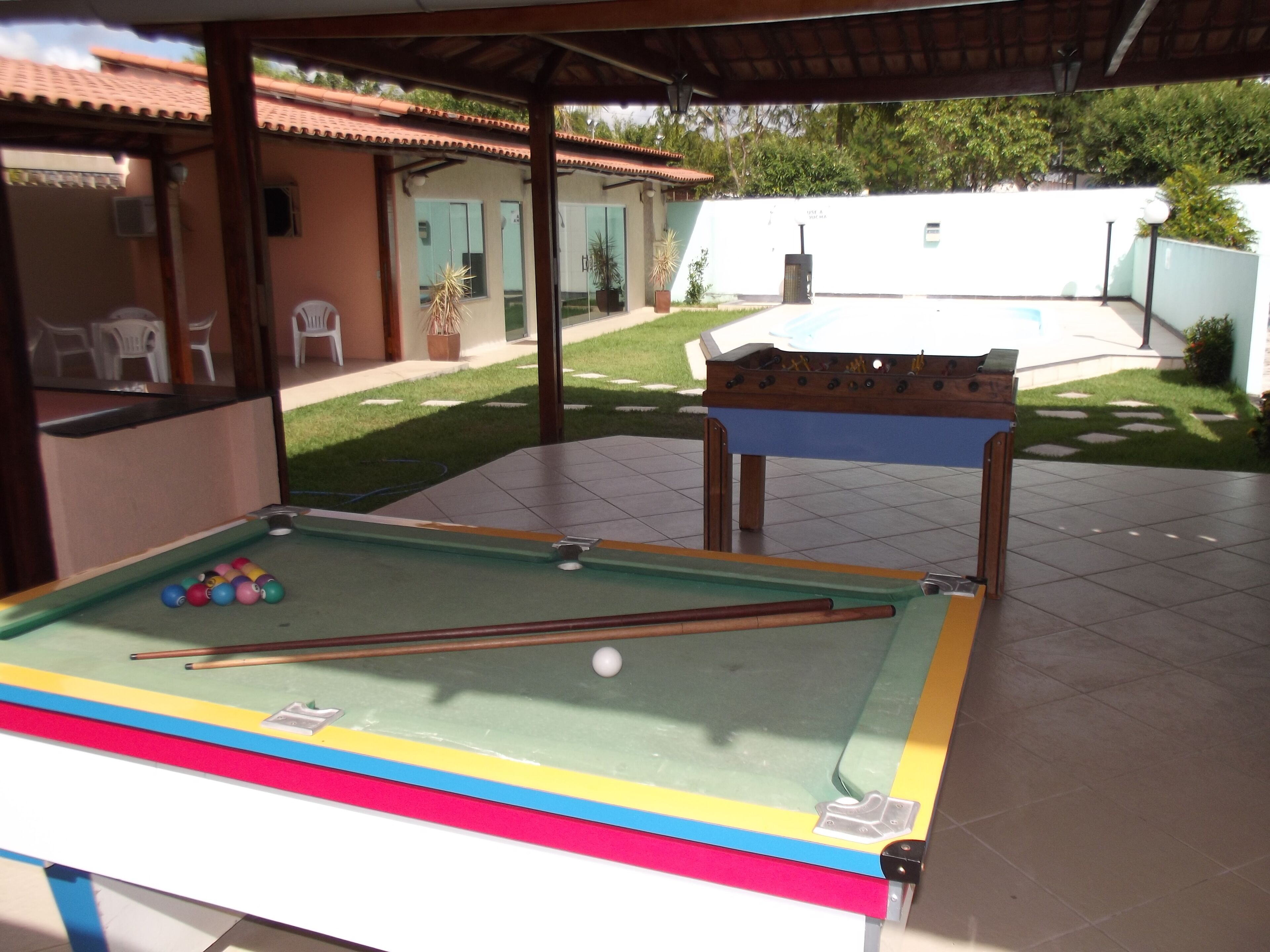 Mesa de bilhar com bolas e tacos, mesa de futebol de mesa, e área de pátio com espaço de estar coberto e vista para uma casa e gramado.