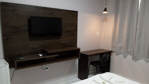 A hotel room with a bed, a desk, a chair, a TV mounted on a wooden panel, and a hanging light fixture. - Hotel Jacarandá (Teixeira de Freitas)