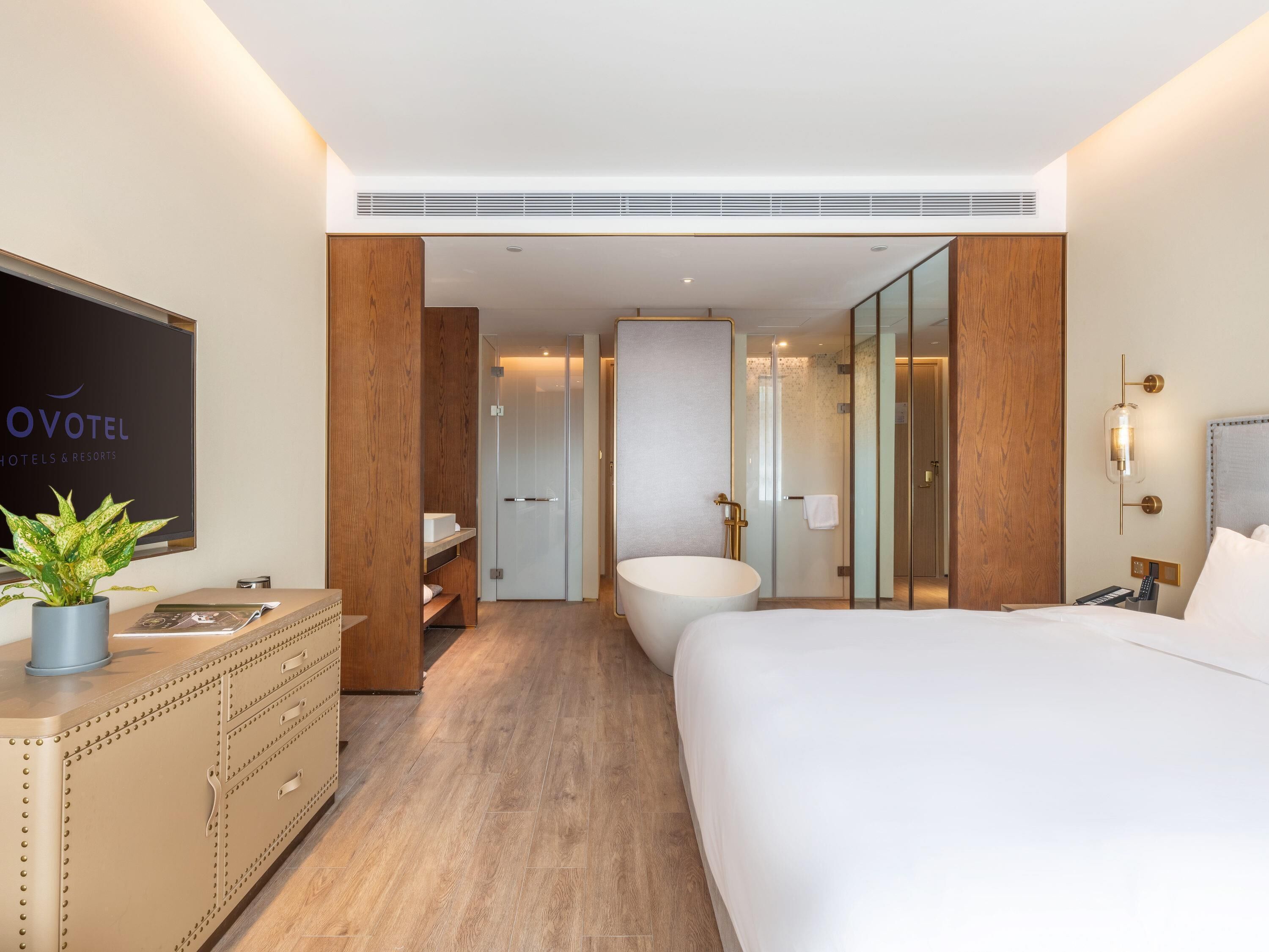 A modern hotel room with a large bed, a bathroom with a bathtub, a wooden dresser, and a television on the wall.