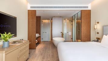 A modern hotel room with a large bed, a bathroom with a bathtub, a wooden dresser, and a television on the wall.