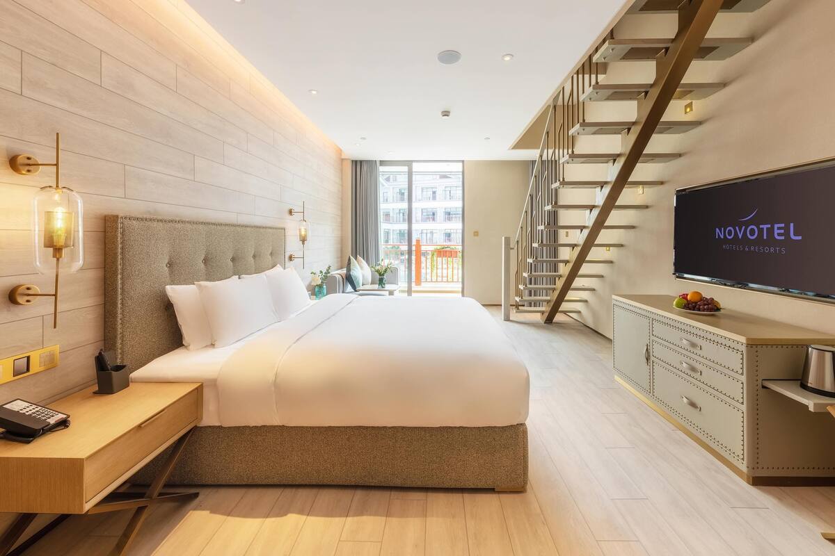 A modern hotel room with a large bed, a desk, a TV, and a staircase.