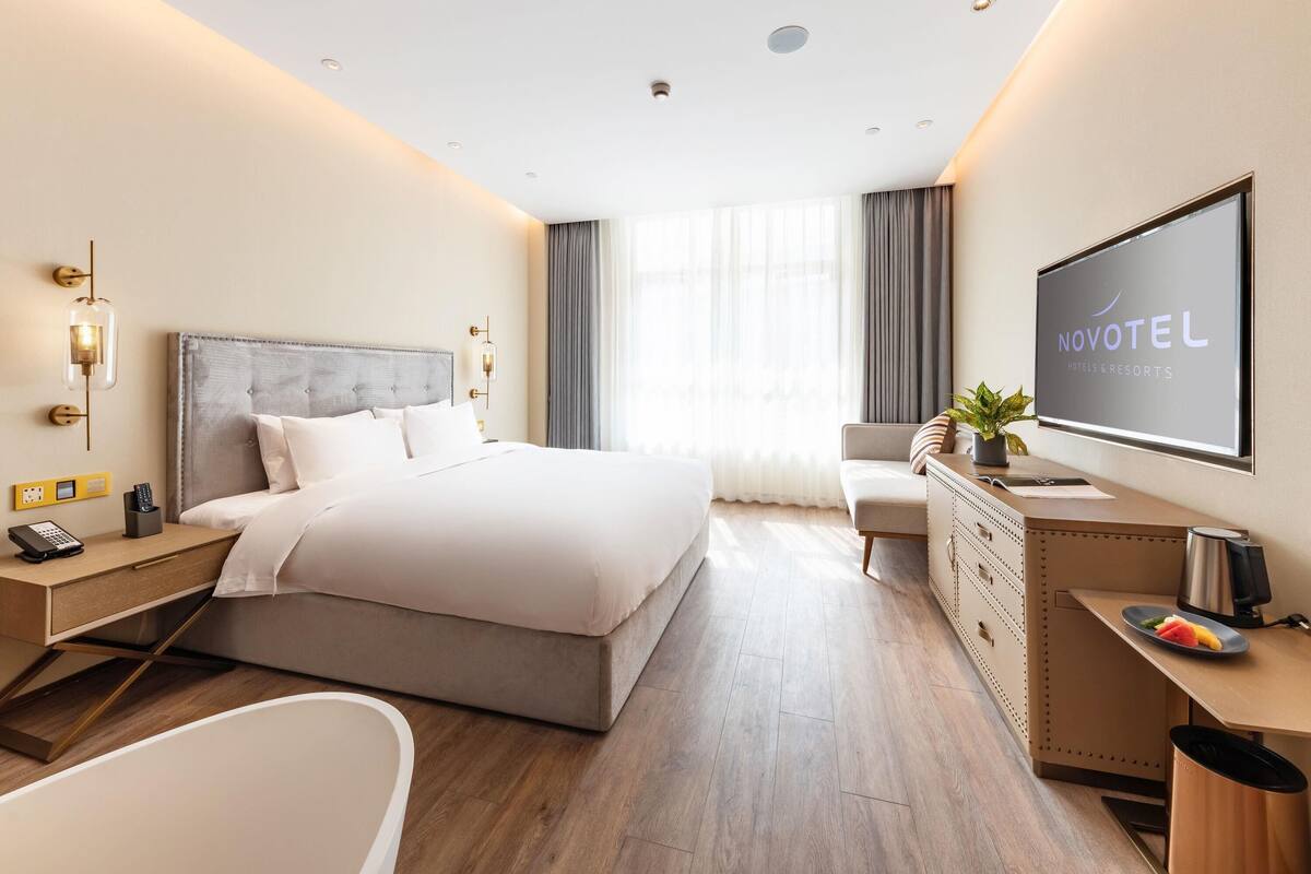 A modern hotel room with a large bed, a desk, a chair, a desk lamp, a TV, and a bathroom with a bathtub.