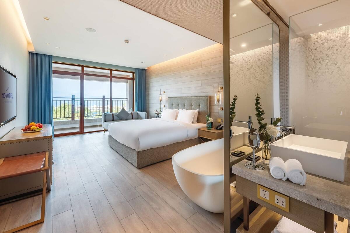 A modern hotel room with a large bed, a separate bathroom with a bathtub, a desk with a TV, and a balcony with a view.