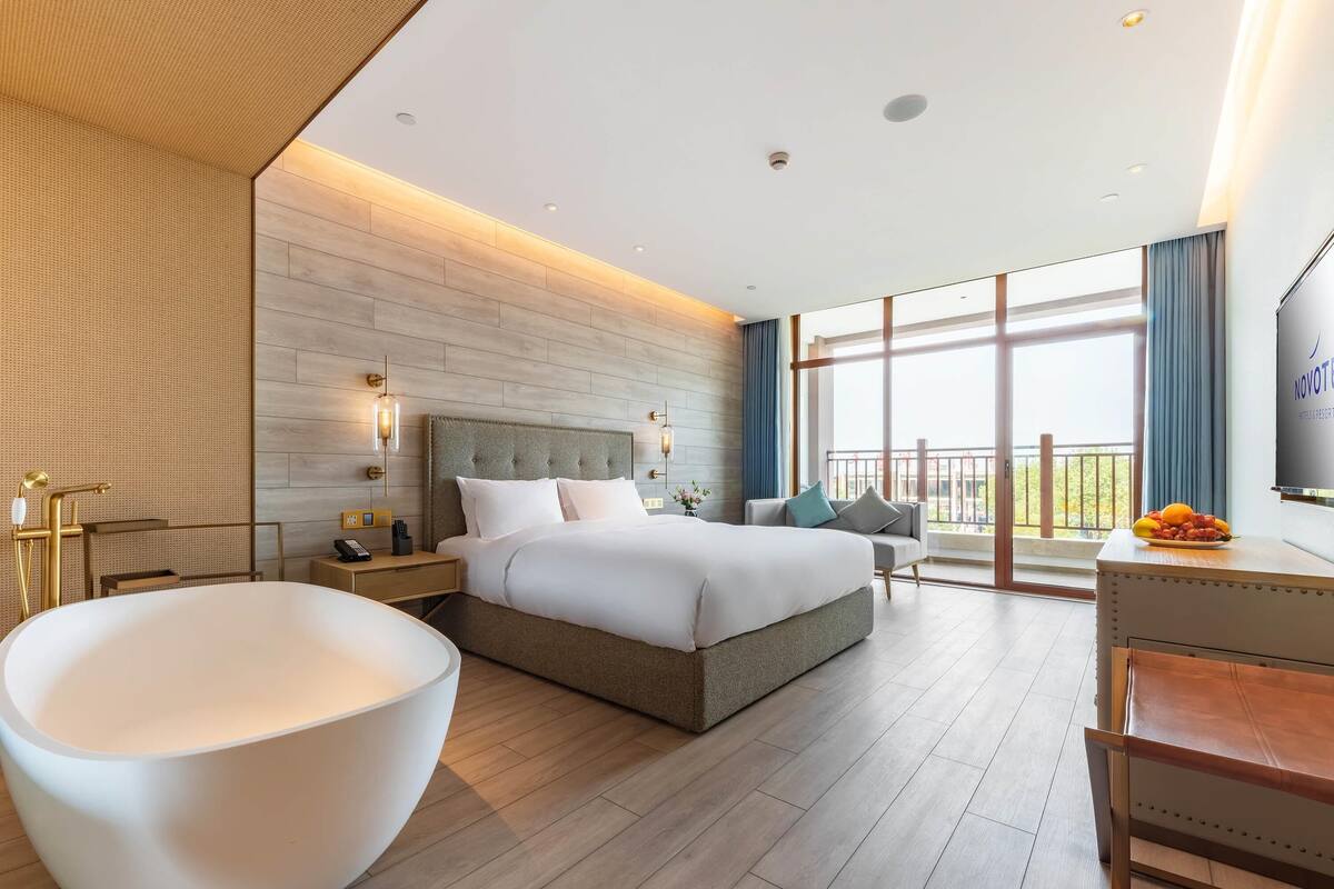 A modern hotel room with a large bed, a sitting area with a sofa, a large white bowl, and a balcony with a view.