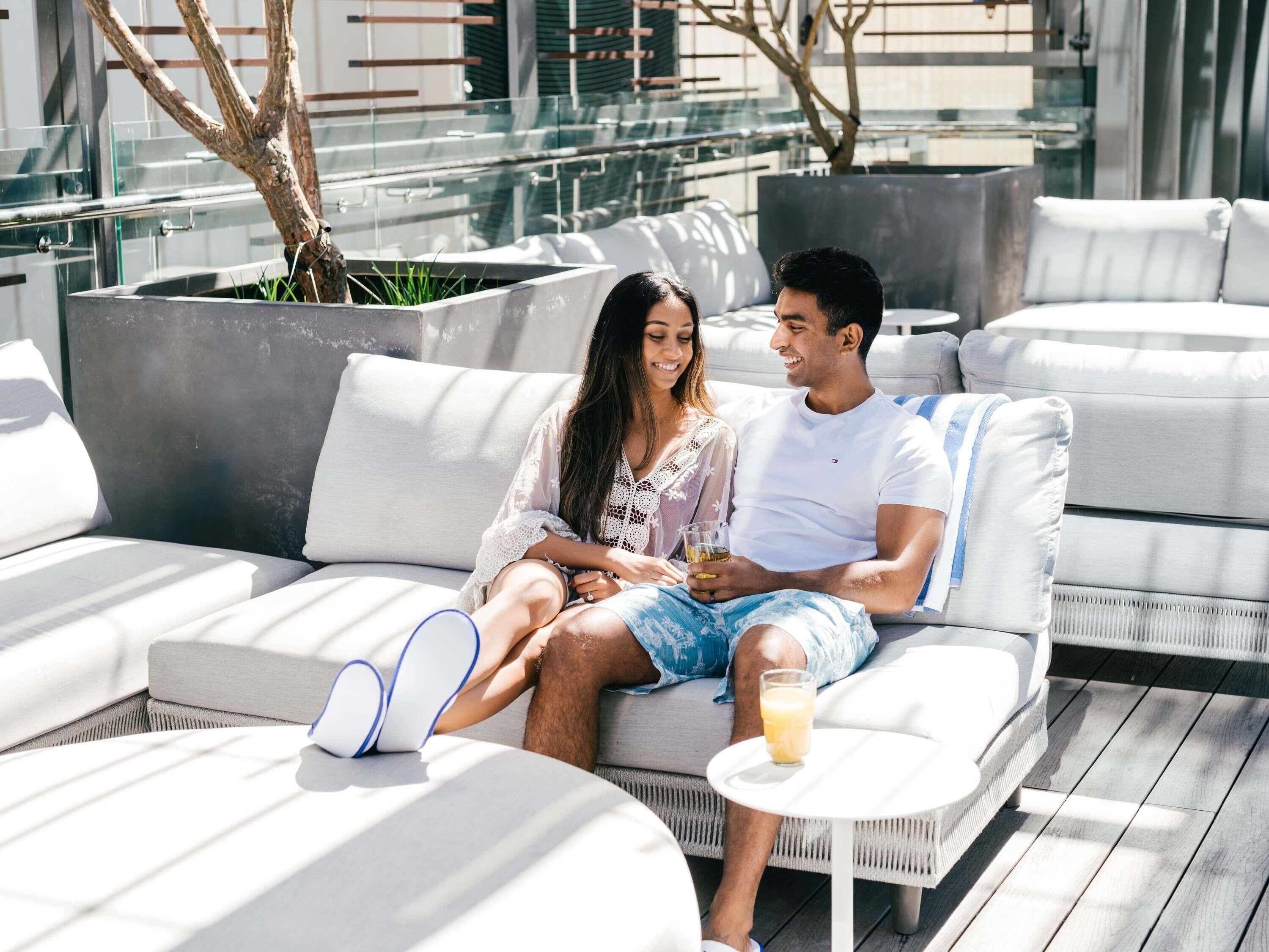 A couple sitting on a white outdoor sofa with a glass of drink on a small table beside them.
