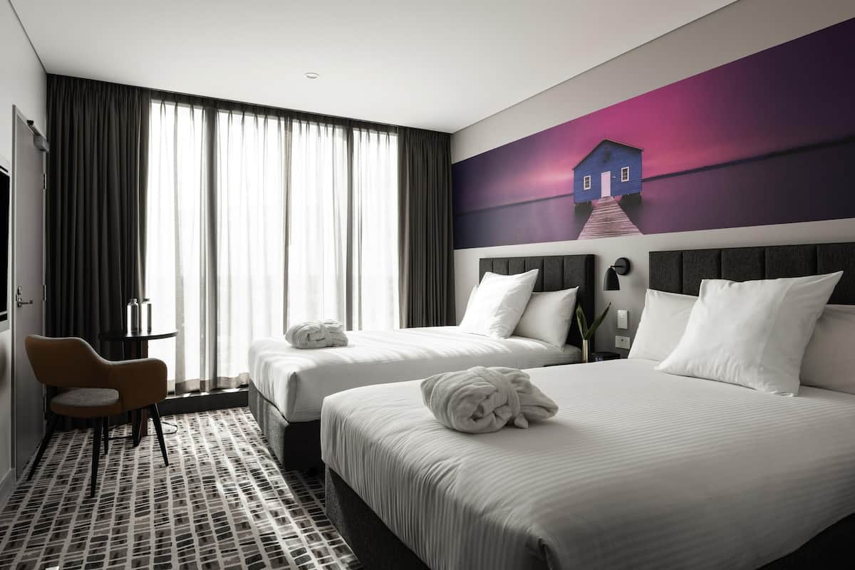 Habitación de hotel con dos camas, un escritorio, una silla y un gran ventanal con cortinas.