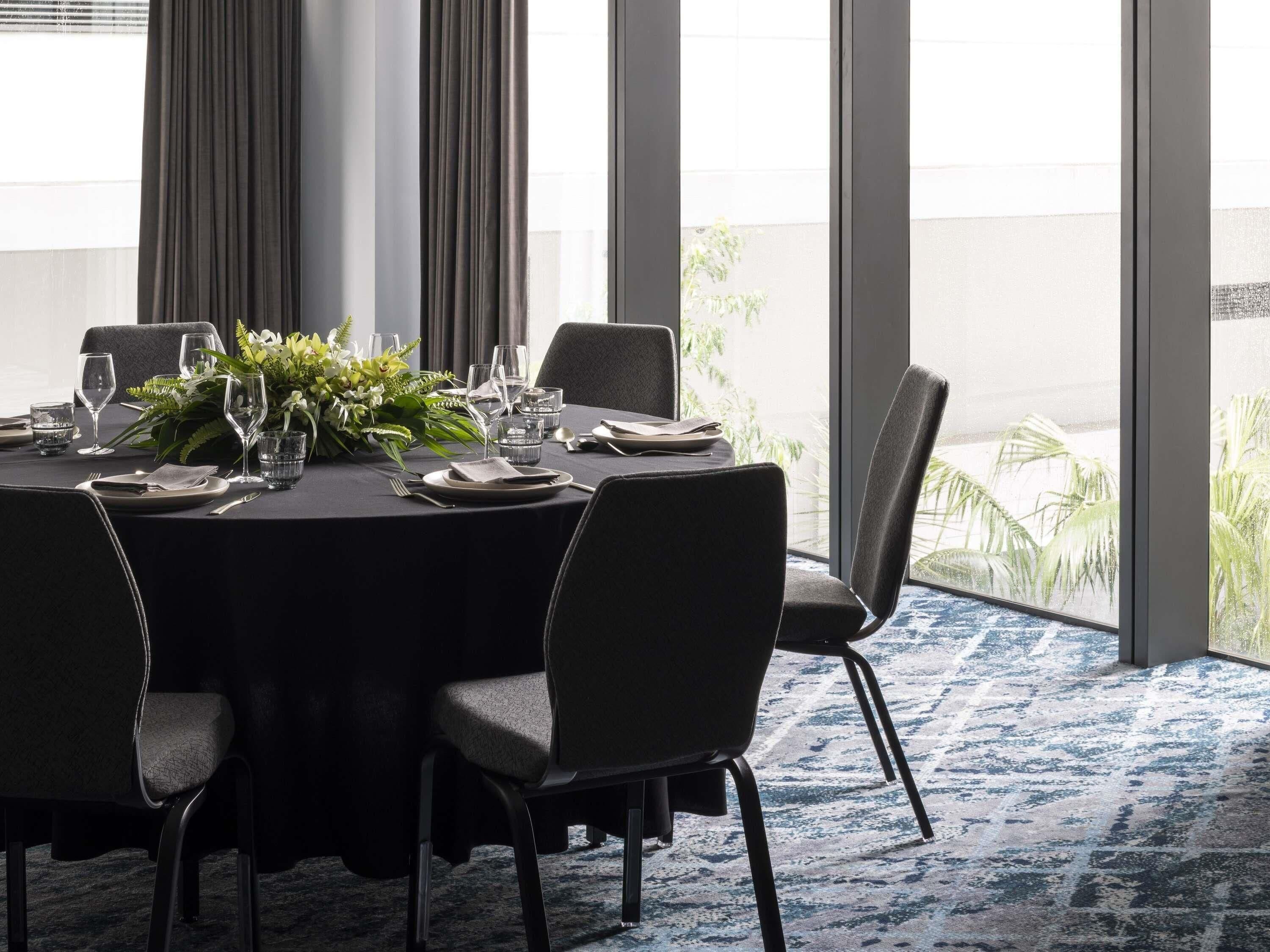 A dining area with a round table set for four, surrounded by chairs, with a large window and curtains.