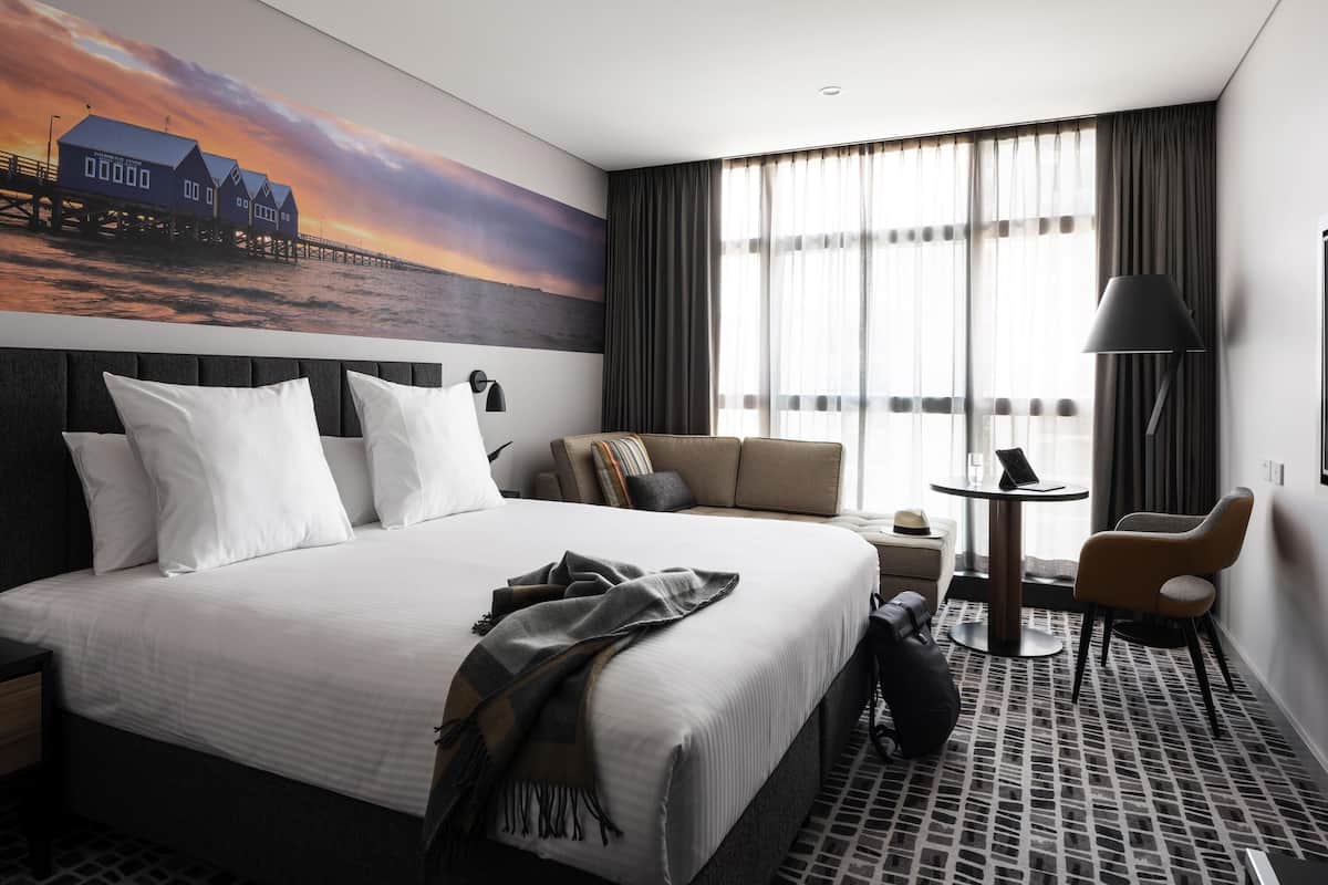 A hotel room with a large bed, a sofa, a small table, and a chair. There is a painting of a pier on the wall.