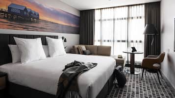 A hotel room with a large bed, a sofa, a small table, and a chair. There is a painting of a pier on the wall.