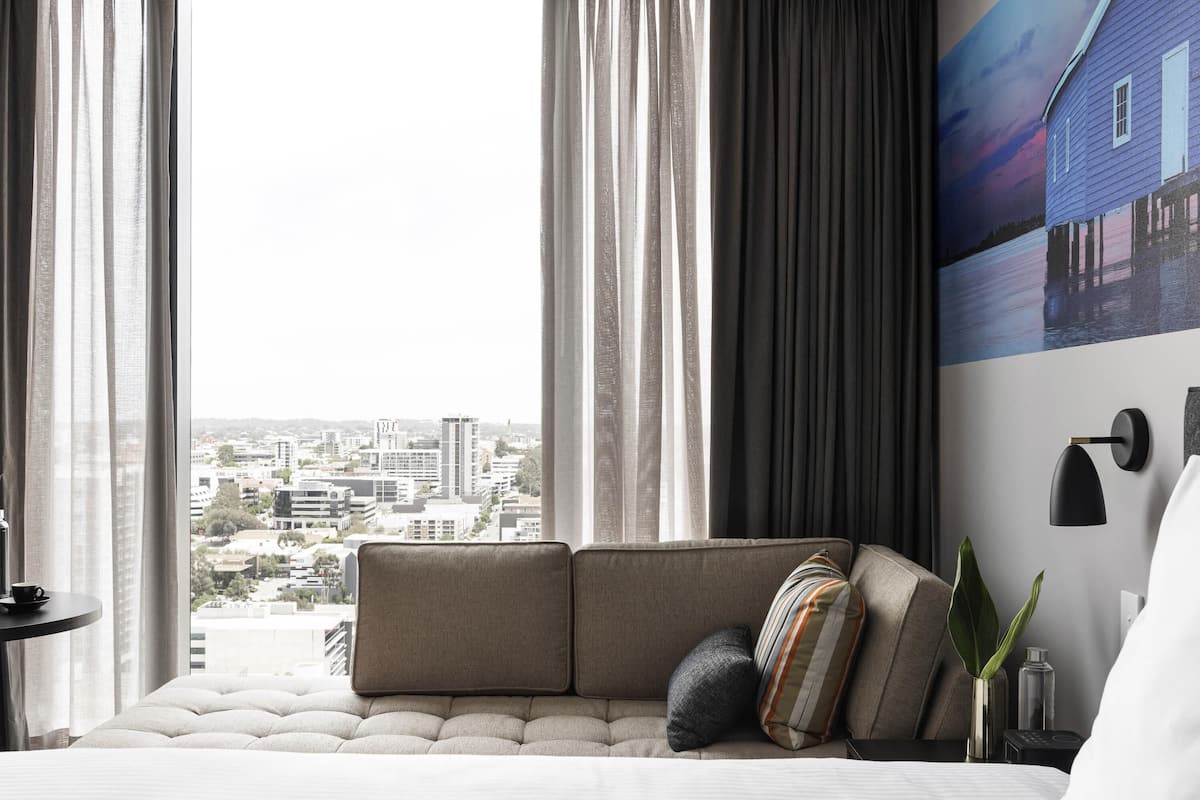 A modern hotel room with a large window, a sofa, a bedside table, and a wall-mounted lamp.
