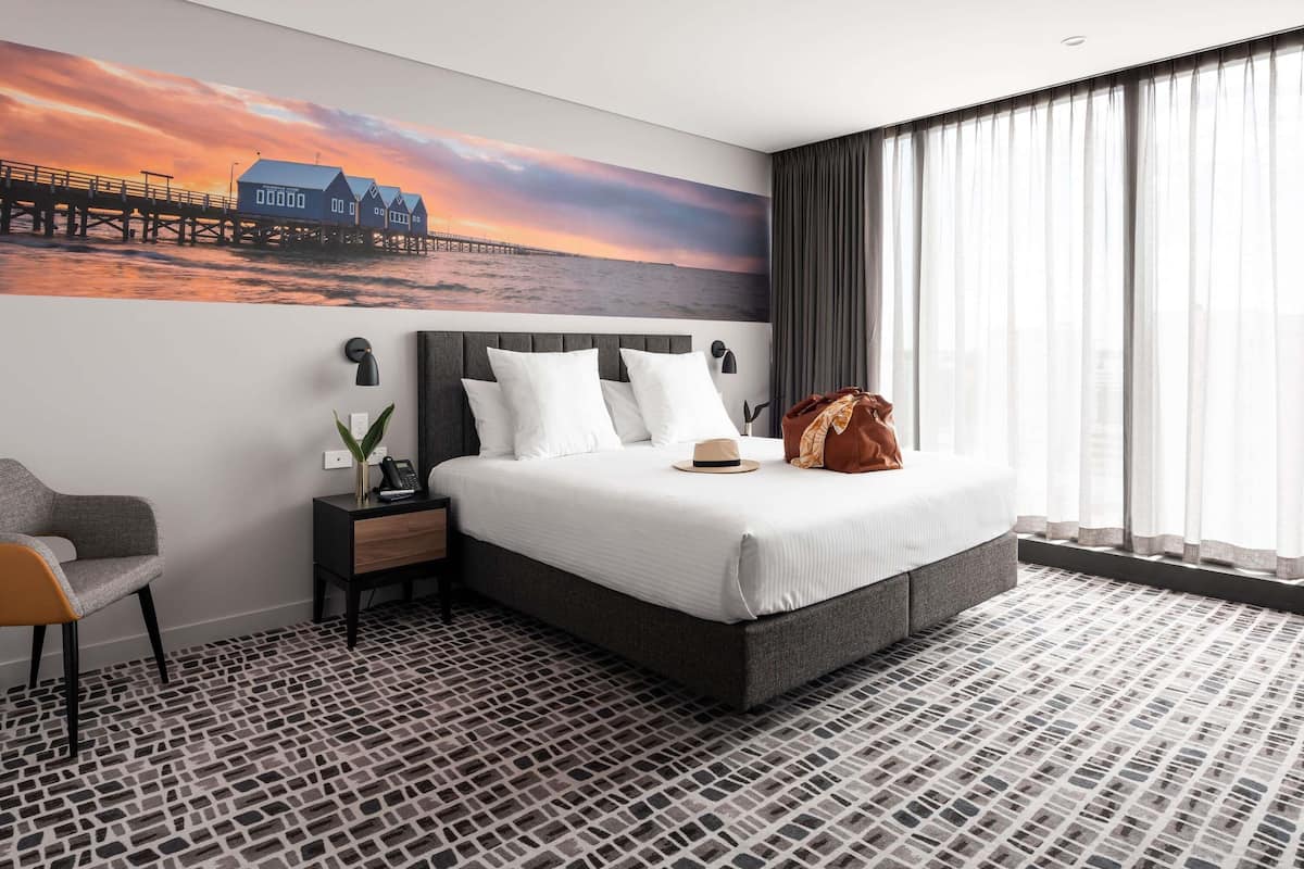 A hotel room with a large bed, a nightstand, a chair, a wall-mounted lamp, and a view of a pier at sunset.