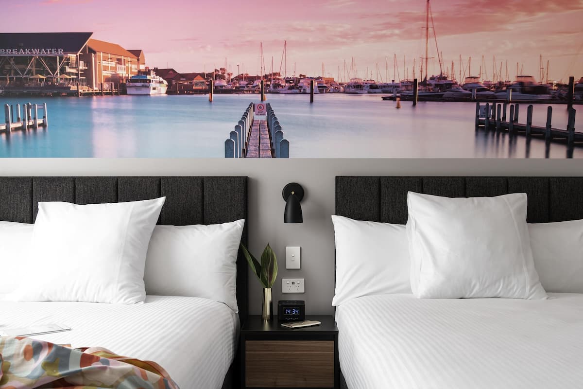 A hotel room with two beds, a nightstand with a clock, a wall-mounted lamp, and a view of a marina with boats.
