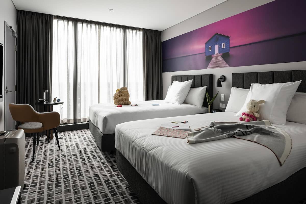 A hotel room with two beds, a desk, a chair, and a large window with curtains.