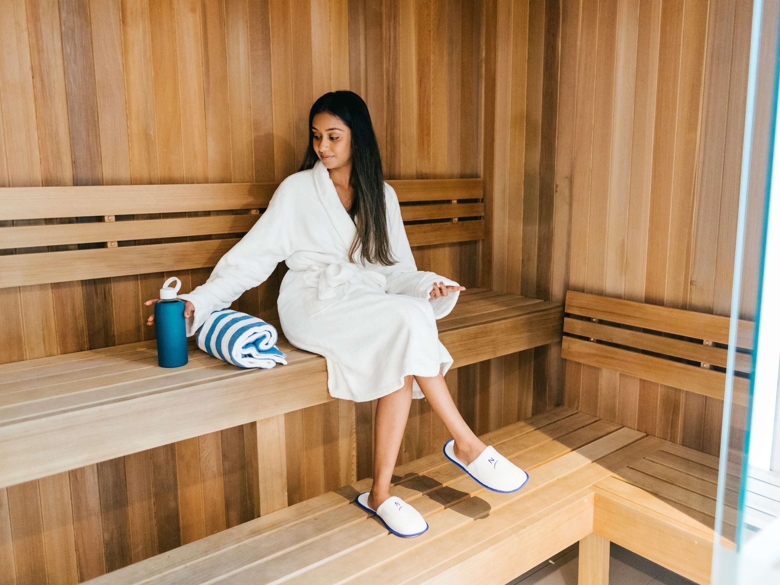 A person sitting on a wooden bench in a sauna area with a towel and a water bottle beside them.