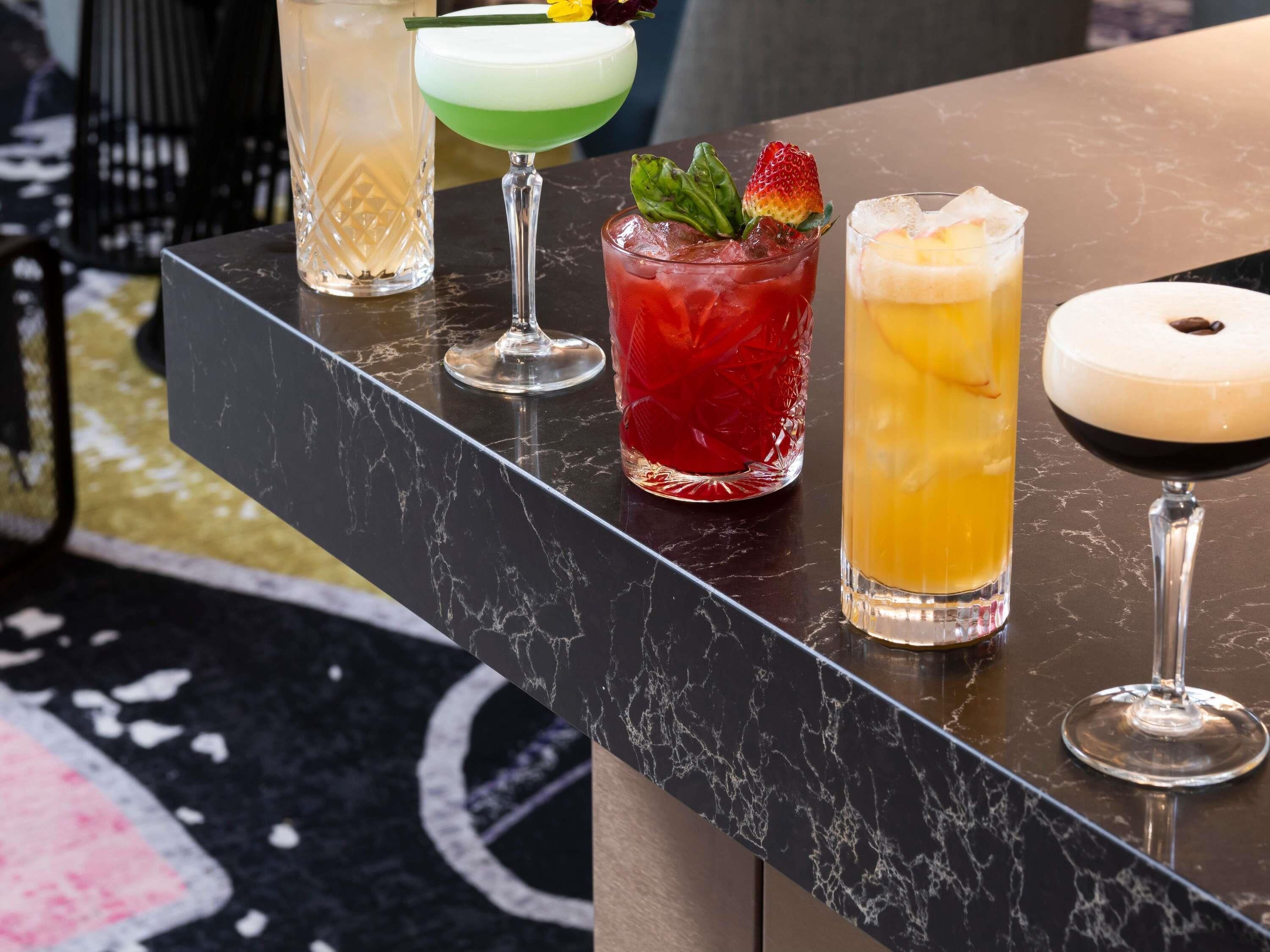 Un bancone bar con diverse bevande, tra cui un cocktail verde, una bibita rossa con una fragola e una bevanda gialla con ghiaccio.