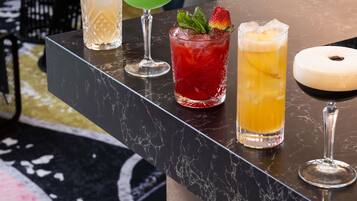 Un bancone bar con diverse bevande, tra cui un cocktail verde, una bibita rossa con una fragola e una bevanda gialla con ghiaccio.