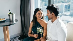 A couple seated on a couch with a city view, a table with a bottle, and a lamp in a room with a modern design. - Novotel Perth Murray Street (Perth)