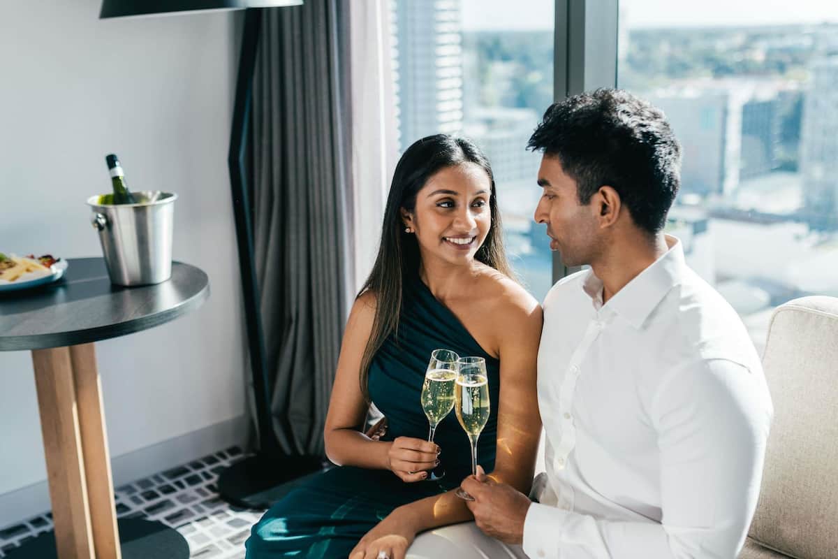 A couple seated on a couch with a city view, a table with a bottle, and a lamp in a room with a modern design.