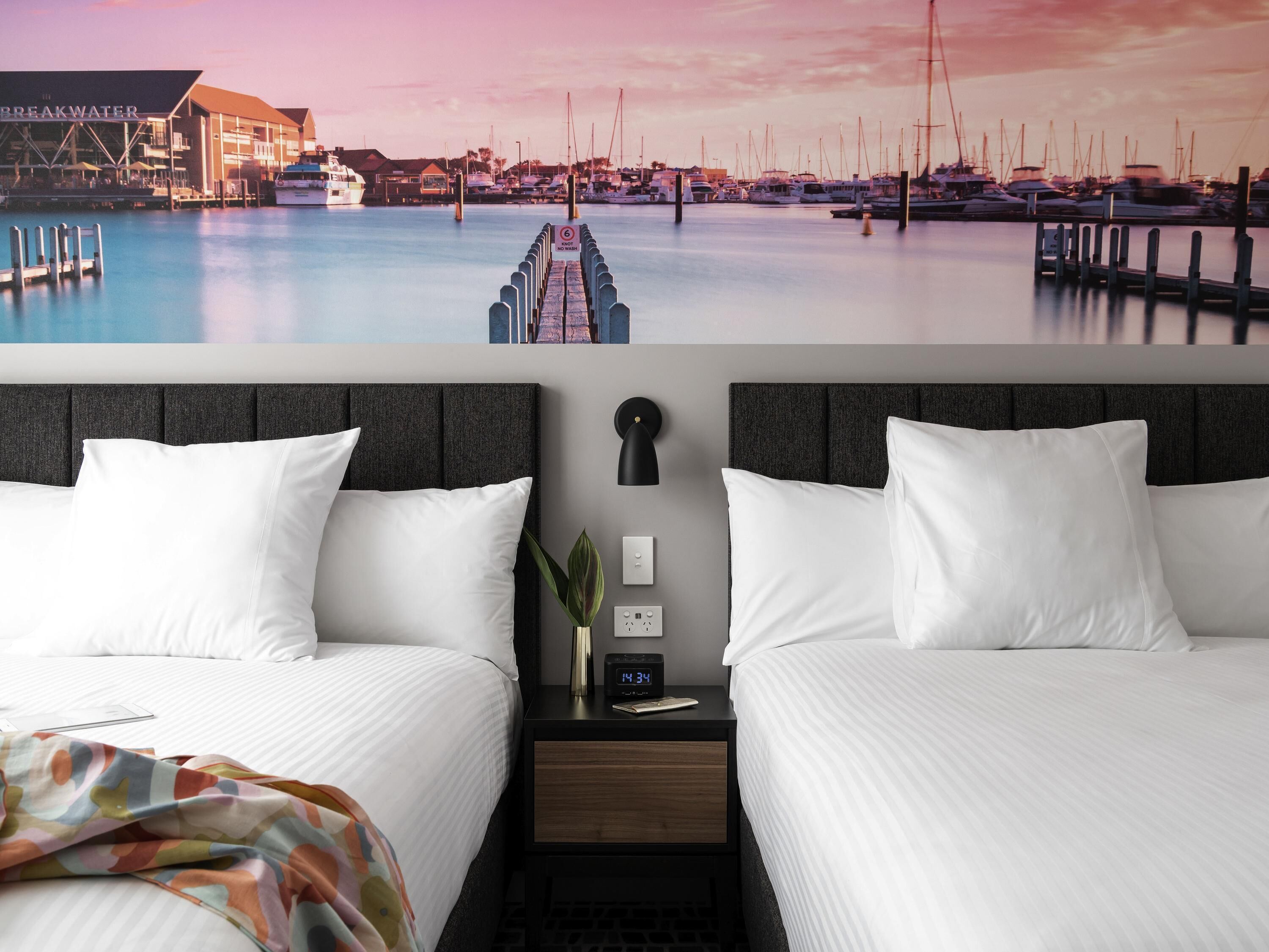 A hotel room with two beds, a nightstand with a clock, a wall-mounted lamp, and a view of a marina with boats.