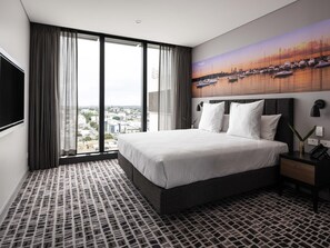 A hotel room with a large bed, a city view, a painting of boats, and a patterned floor.