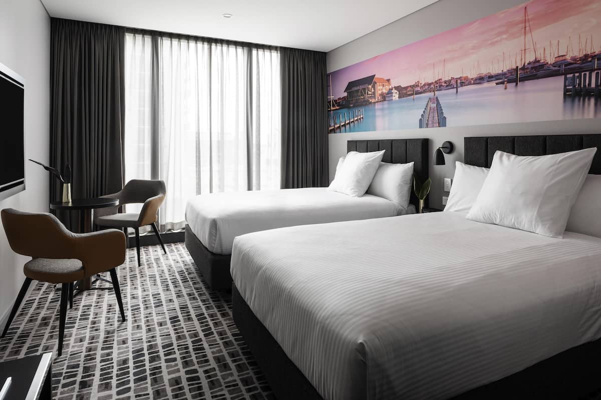 A hotel room with two beds, a desk, chairs, a large window with curtains, and a cityscape mural on the wall.