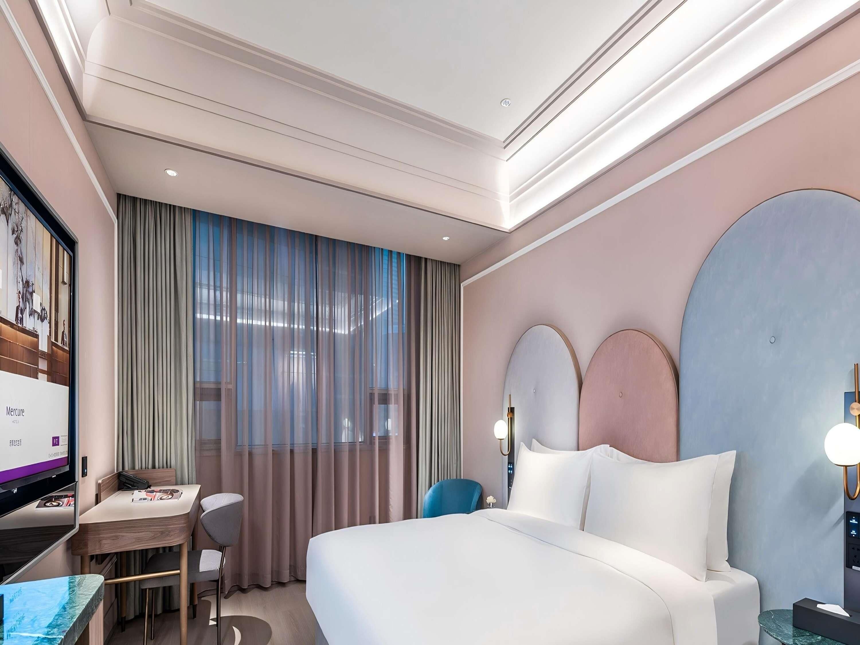 Habitación de hotel con cama, escritorio, silla, televisor y ventana con cortinas.