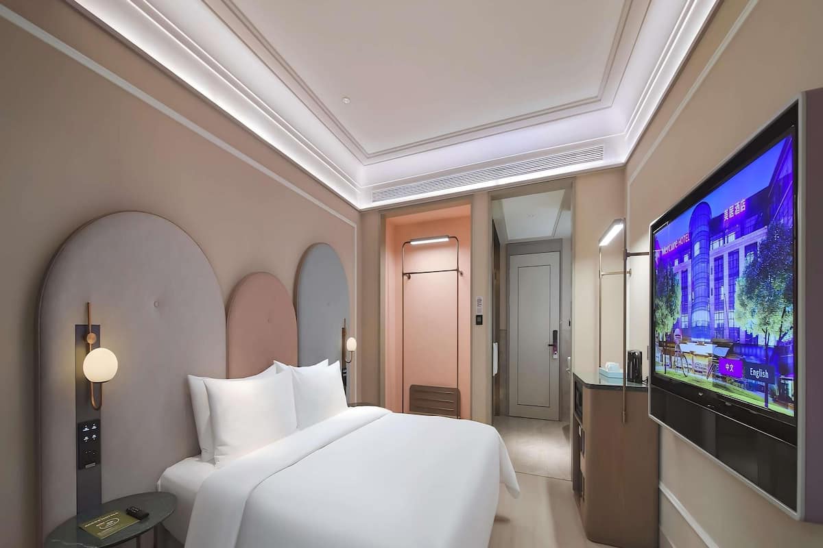 Habitación de hotel con cama, televisión, televisor montado en la pared, una puerta y una mesita con el control remoto.