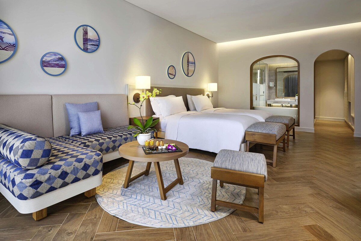 A hotel room with a sofa, two beds, a small table, and a mirror.