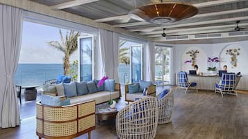 A spacious room with a wooden floor, a sofa, wicker chairs, a desk with two people working, and large windows overlooking the sea.