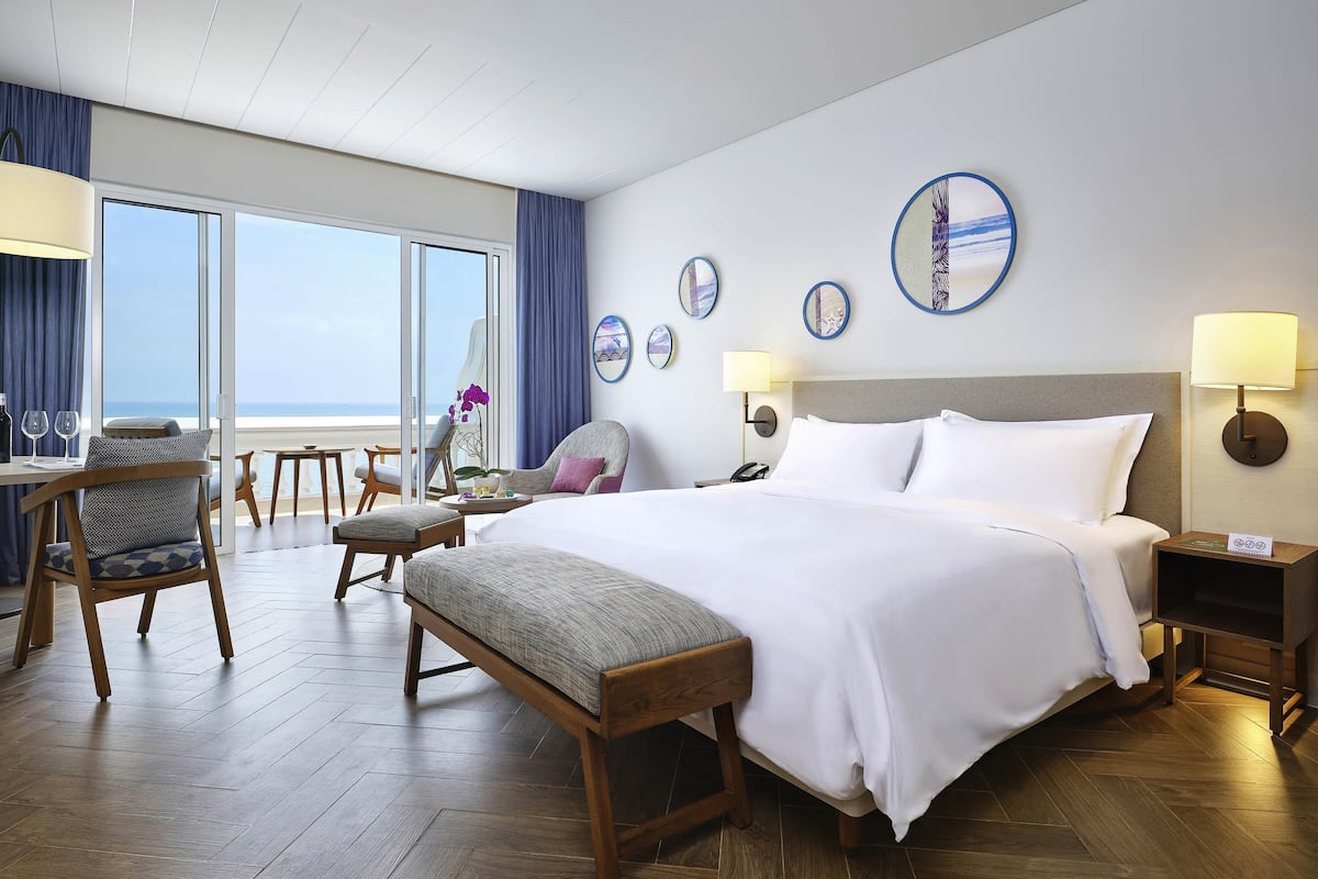 A hotel room with a large bed, a desk with chairs, a view of the ocean, and wall decor.