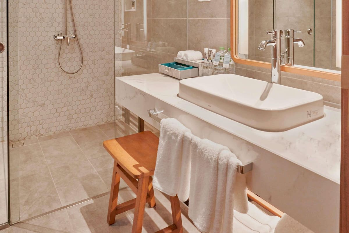 A modern bathroom with a glass shower enclosure, a wooden bench, a white sink, and a mirror above the sink.
