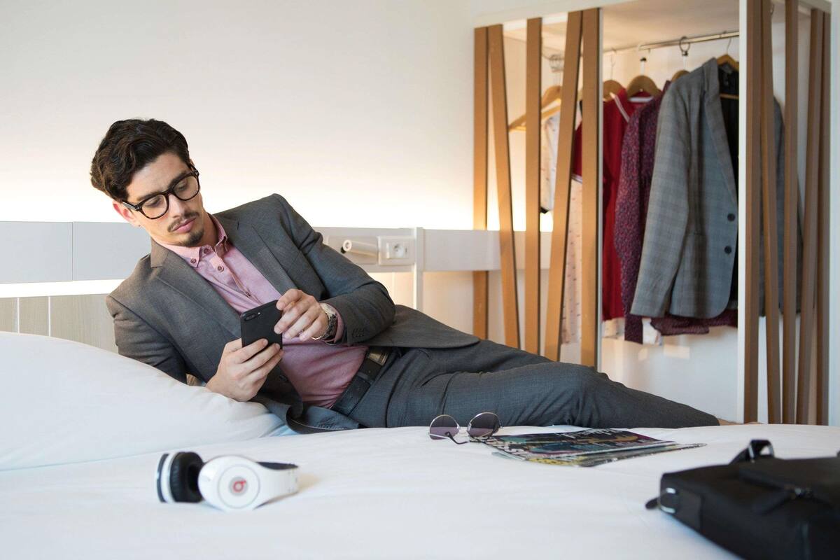 Een man in een pak ligt op een bed in een hotelkamer, met een kledingkast en een tijdschrift op het bed.