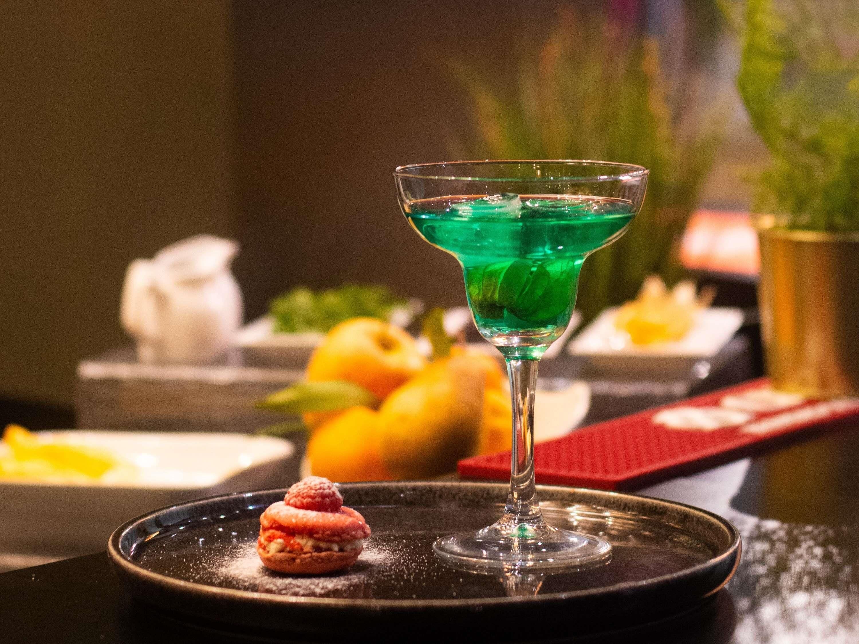 A martini glass with a green liquid, a dessert on a plate, and a blurred background with fruits and a vase.