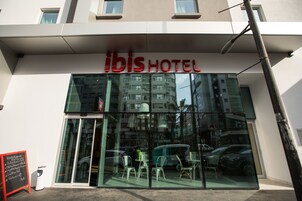 El exterior del hotel ibis con fachada de vidrio, un cartel sobre la entrada y algunas sillas afuera.