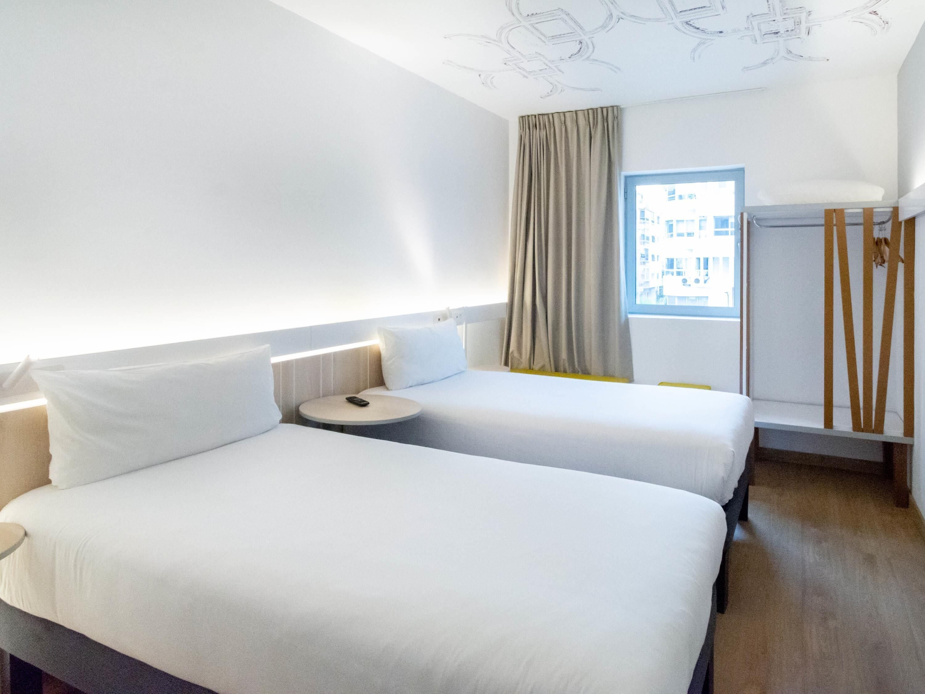 A hotel room with two beds, a small round table, a window with curtains, and a radiator.