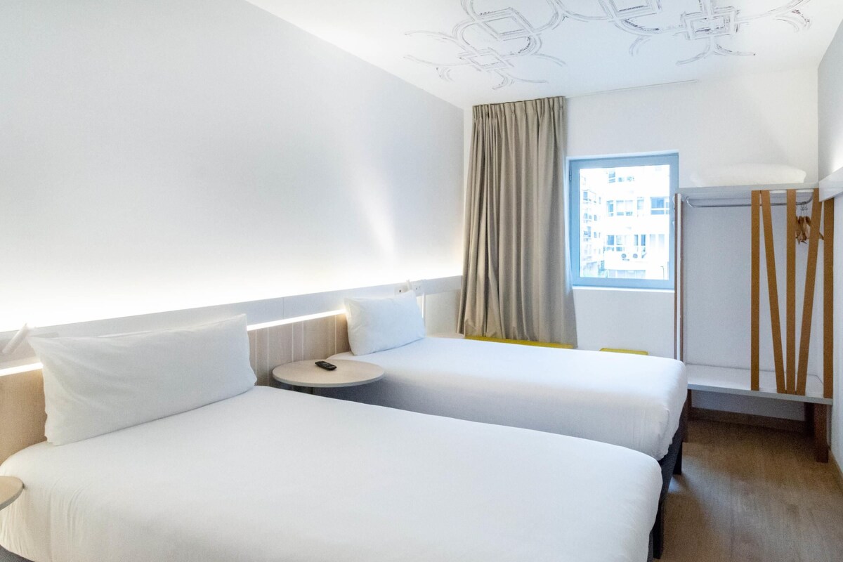 Habitación de hotel con dos camas, una mesita redonda, una ventana con cortinas y un radiador.
