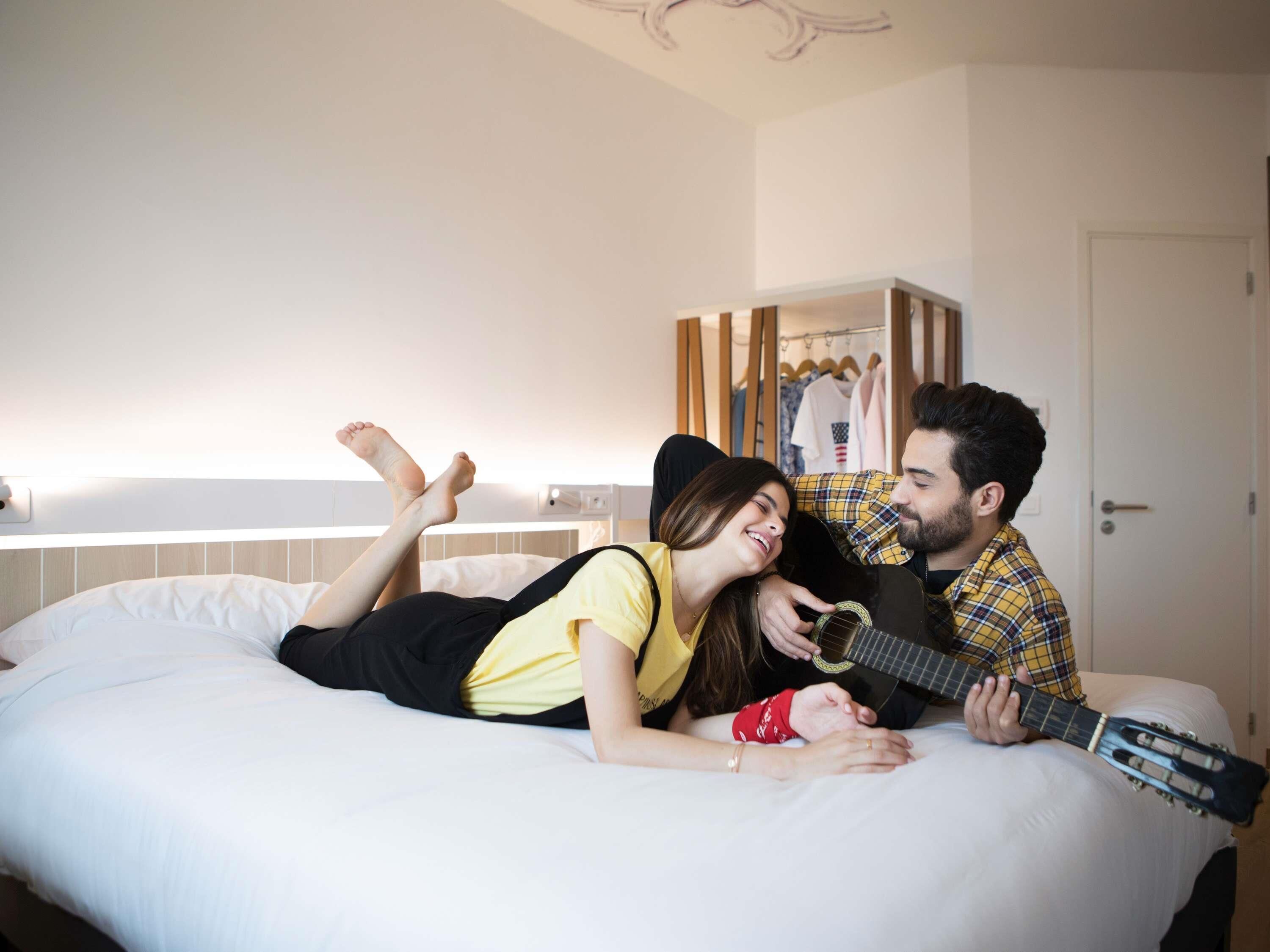 Una pareja acostada en una cama en una habitación de hotel, con un armario y una puerta al fondo.