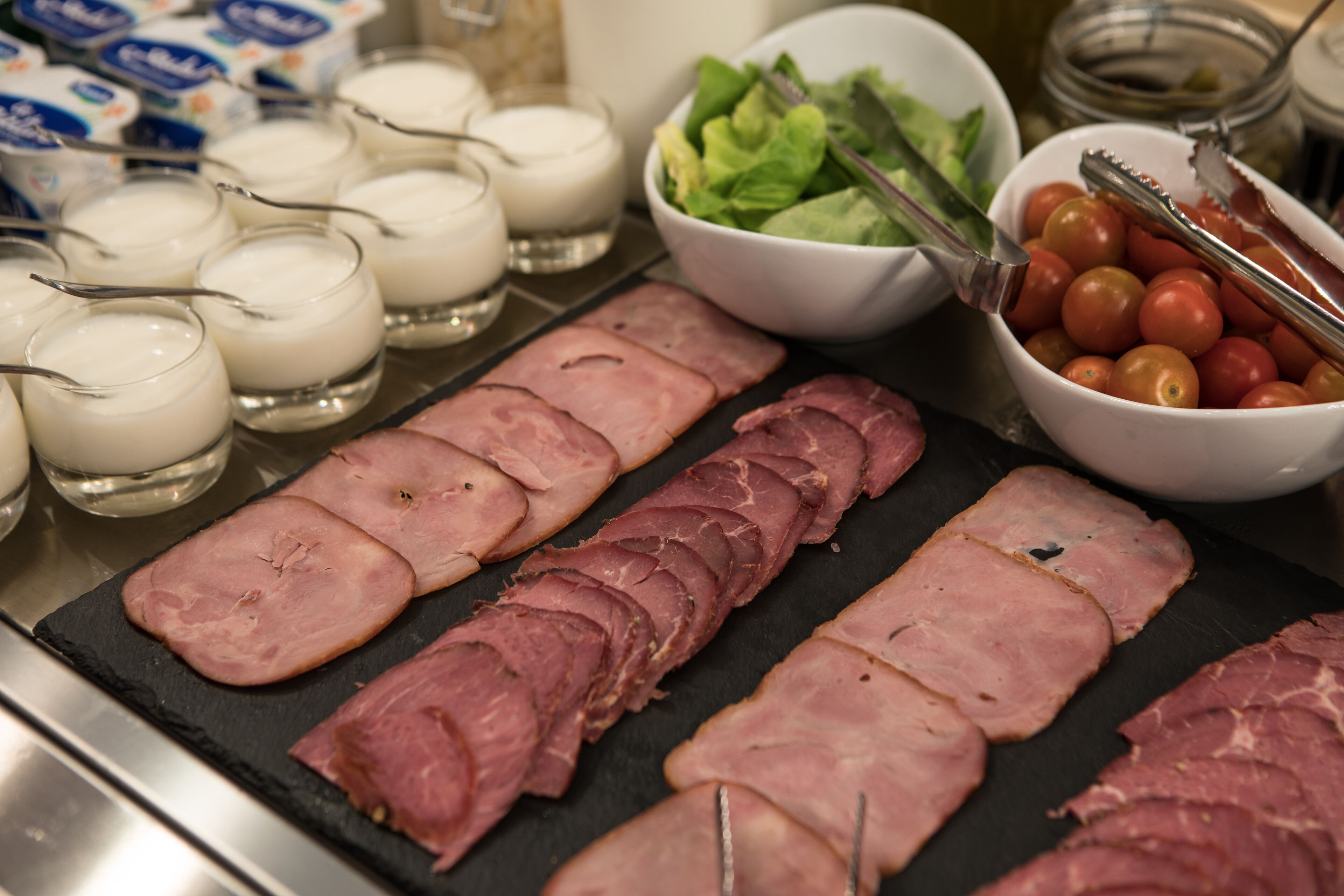 A buffet with sliced ham, a bowl of cherry tomatoes, and a bowl of greens.