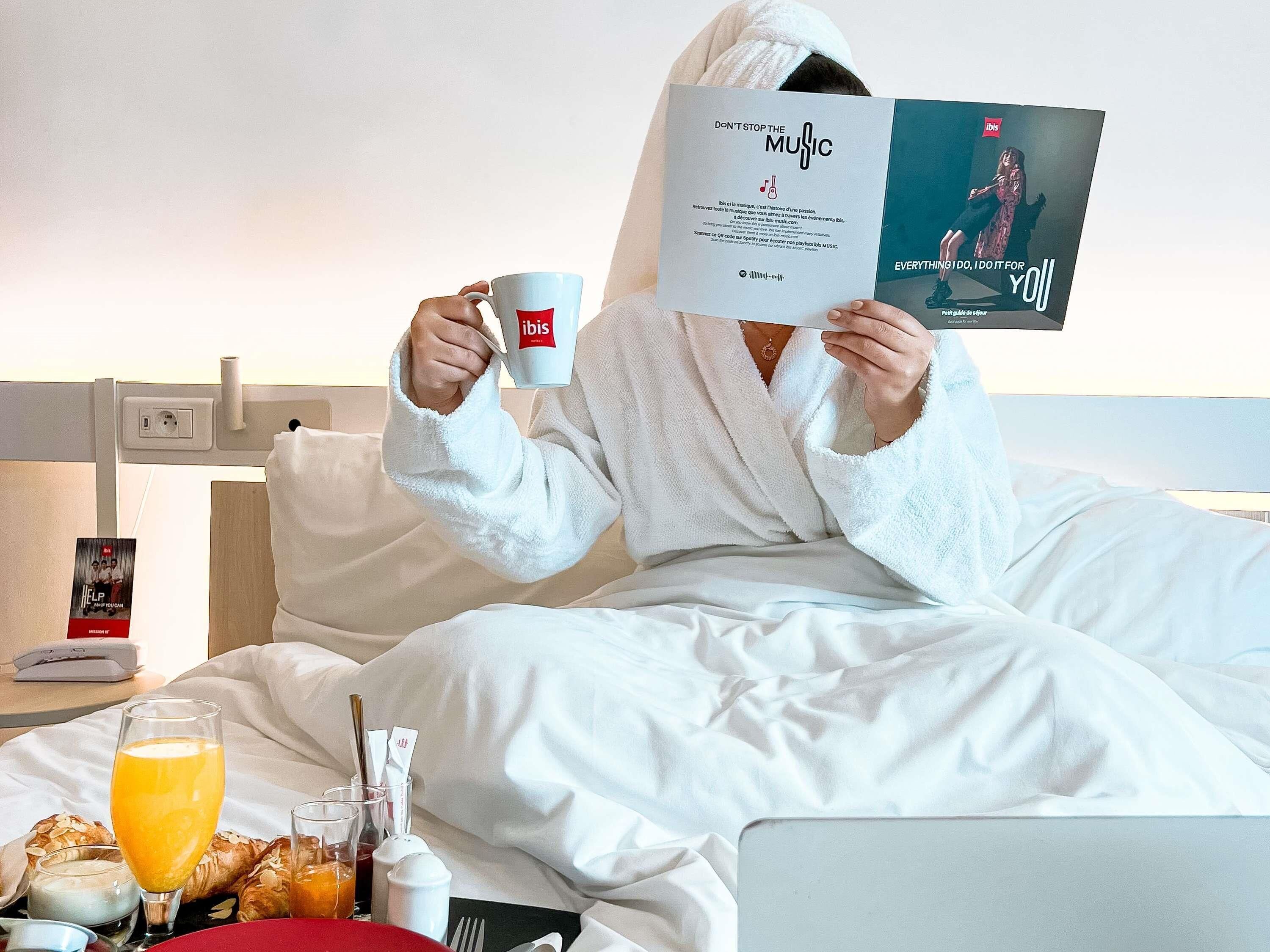 A person in a white bathrobe sitting on a bed with a book and a mug, a laptop, and breakfast items on the bedside table.