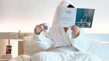 A person in a white bathrobe sitting on a bed with a book and a mug, a laptop, and breakfast items on the bedside table.